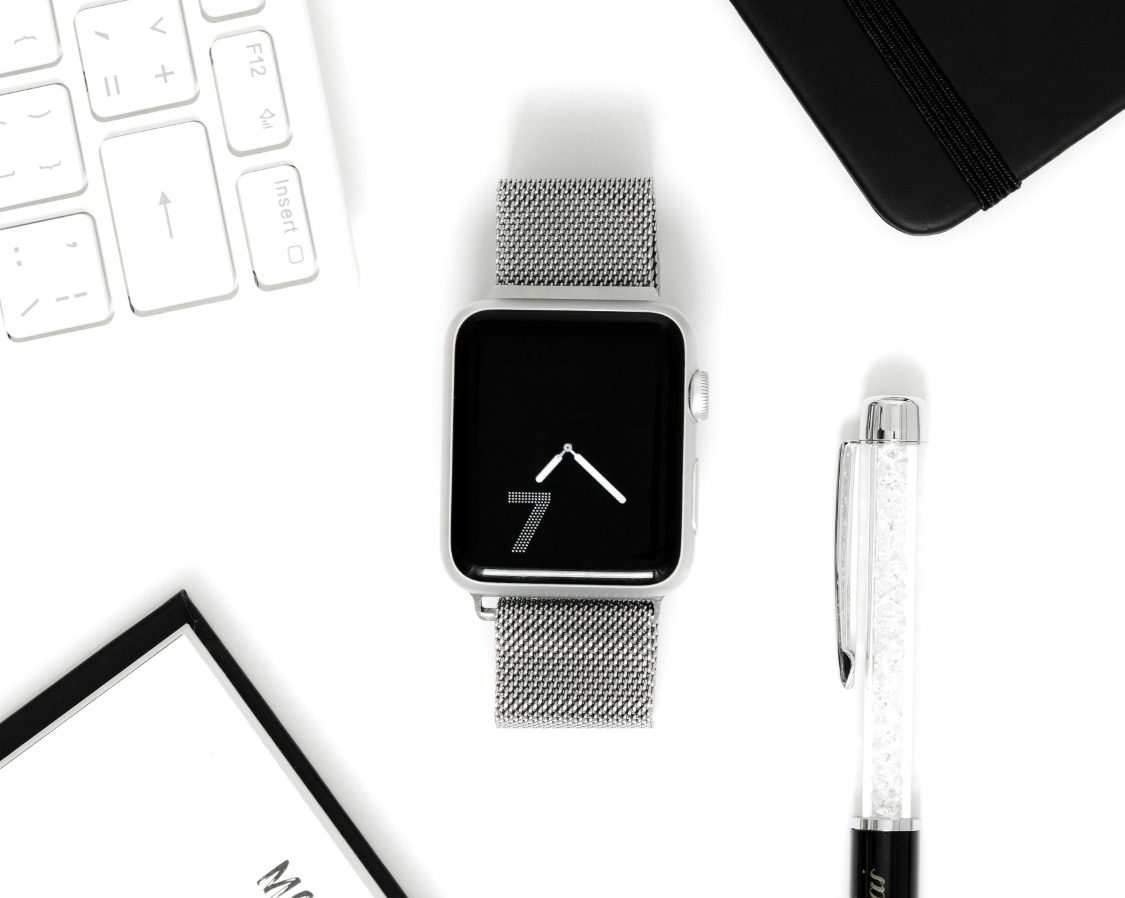 Reloj inteligente con pantalla negra y correa plateada, acompañado de una libreta y bolígrafo.